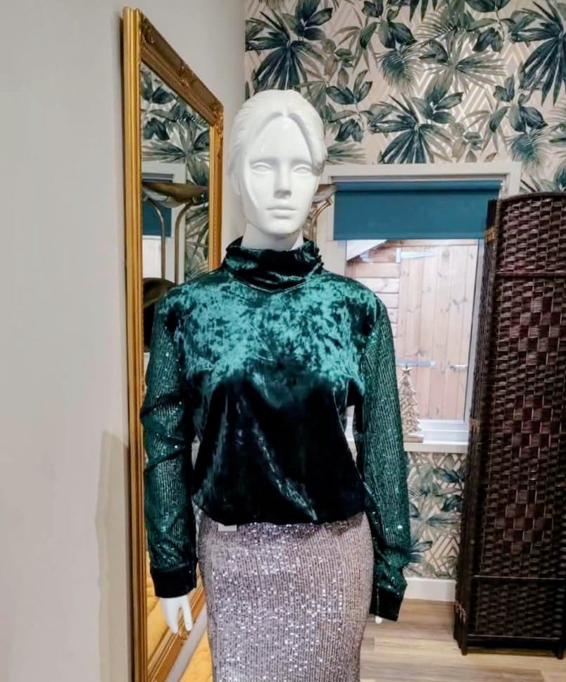 Mannequin wearing a green velvet top and silver skirt in a room with floral wallpaper.