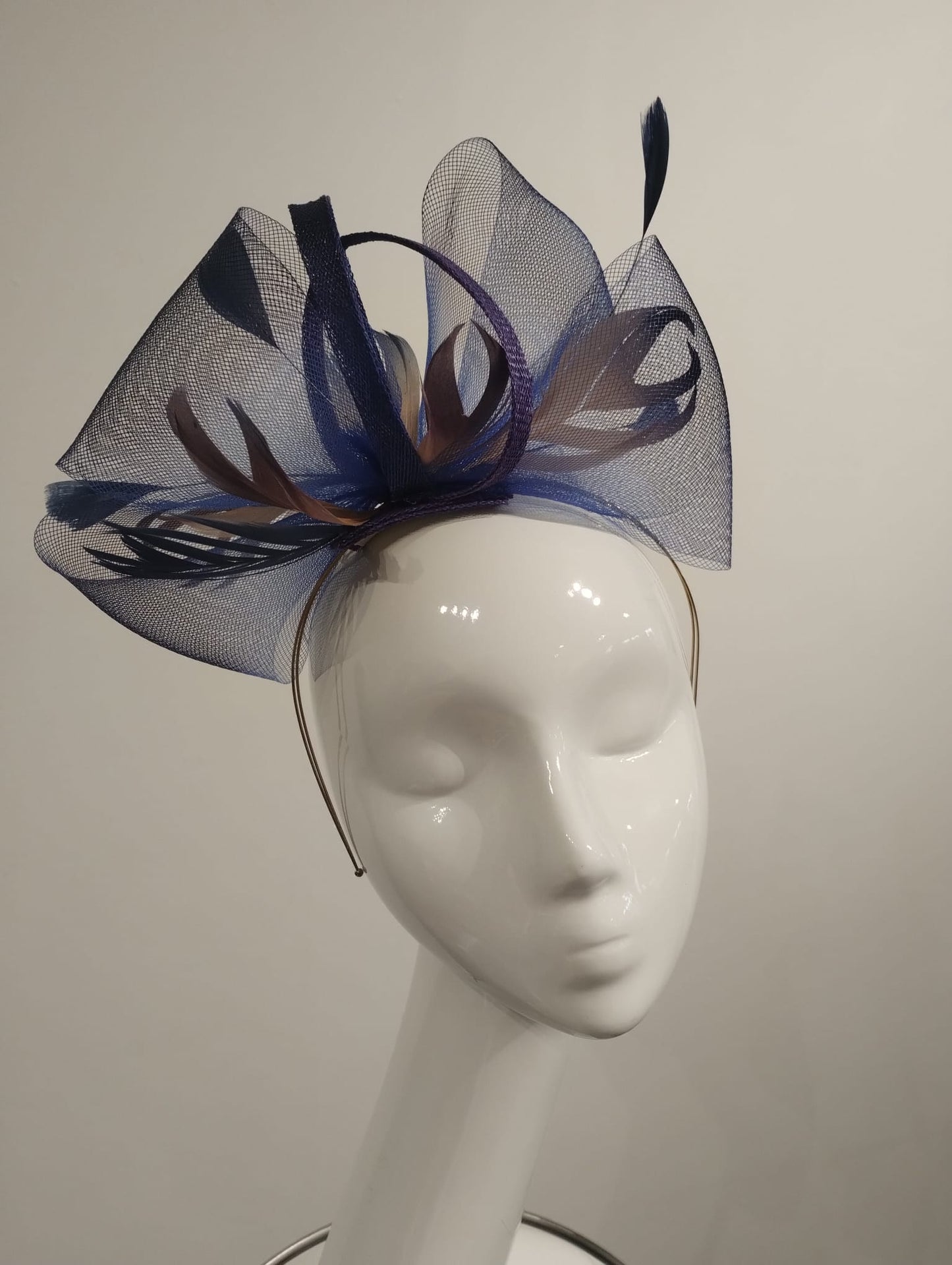 A fascinator with blue and purple feathers and netting on a silver band displayed on a white mannequin head.