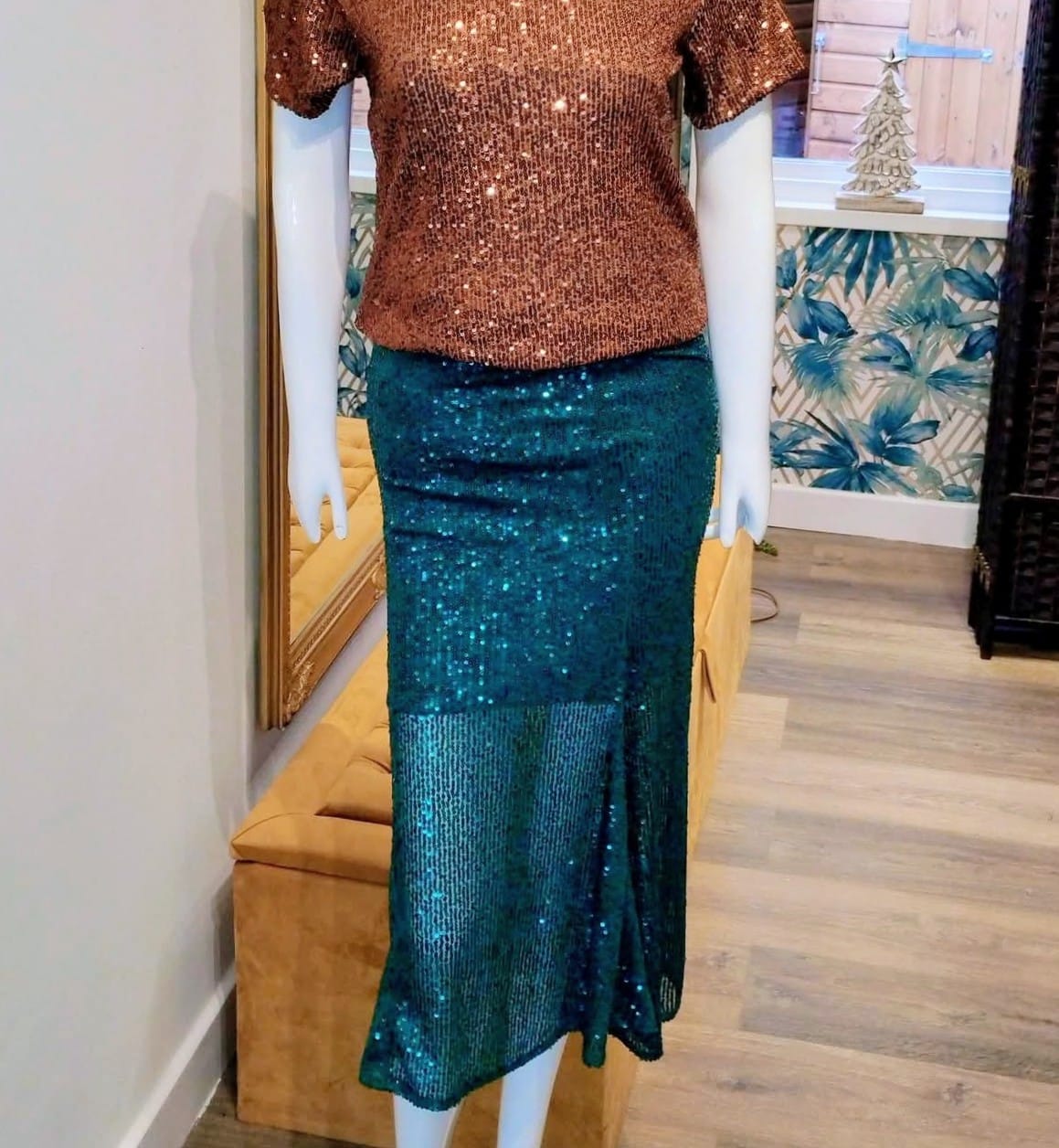 Mannequin wearing a bronze sequin top and teal sequin skirt in a room with a mirror and decorative items.

