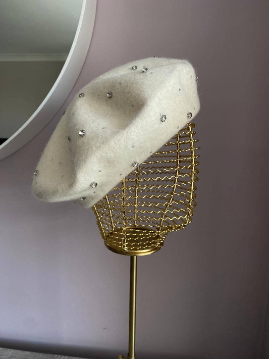 White beret with rhinestones on a gold stand against a neutral background